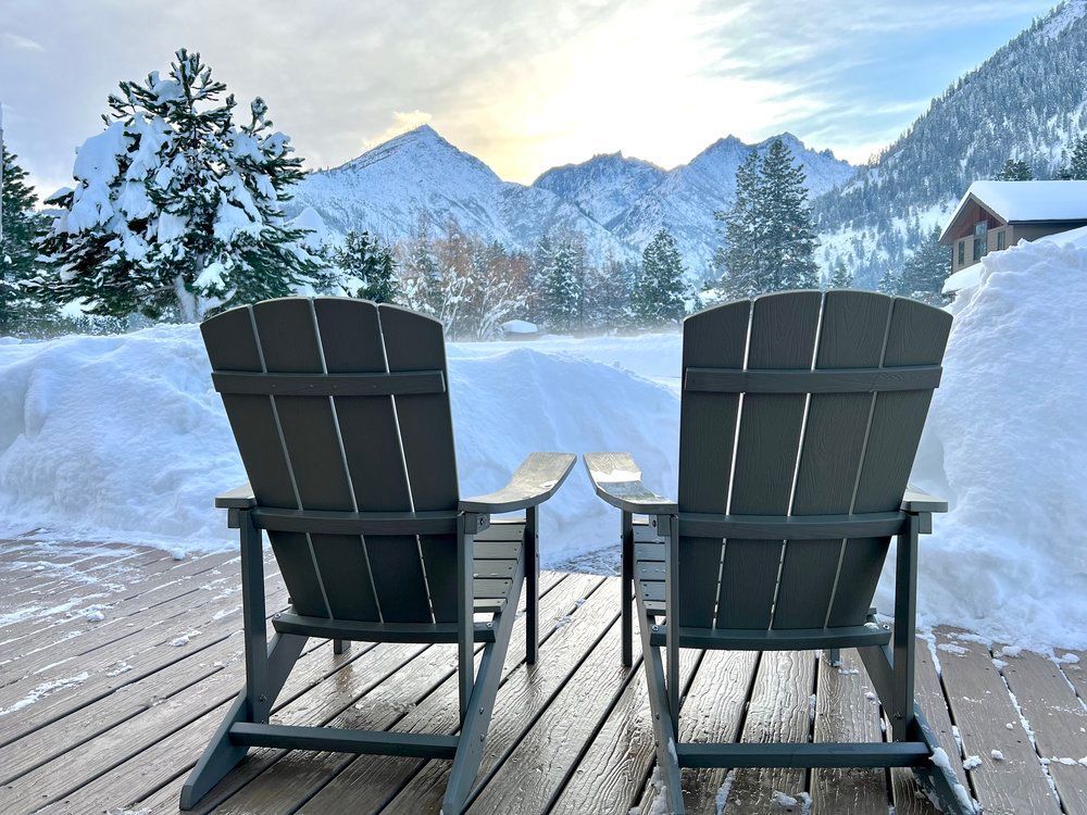Two adirondack chairs sitting on a wooden deck in the snow.