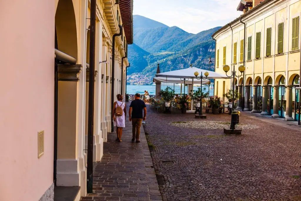 Cobblestone street with people walking between pastel buildings, mountains and lake in the background Cobblestone street with people walking between pastel buildings, mountains and lake in the background