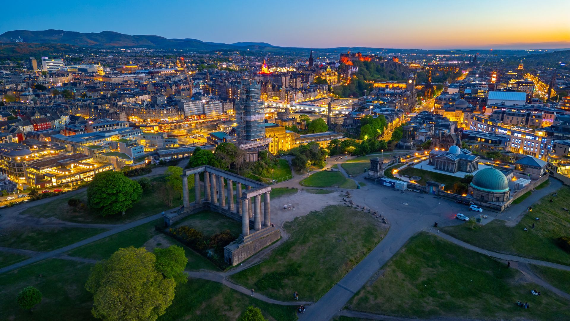 Dusk aerial view of a city skyline with illuminated buildings, a river, and a large stone monument on a grassy hill