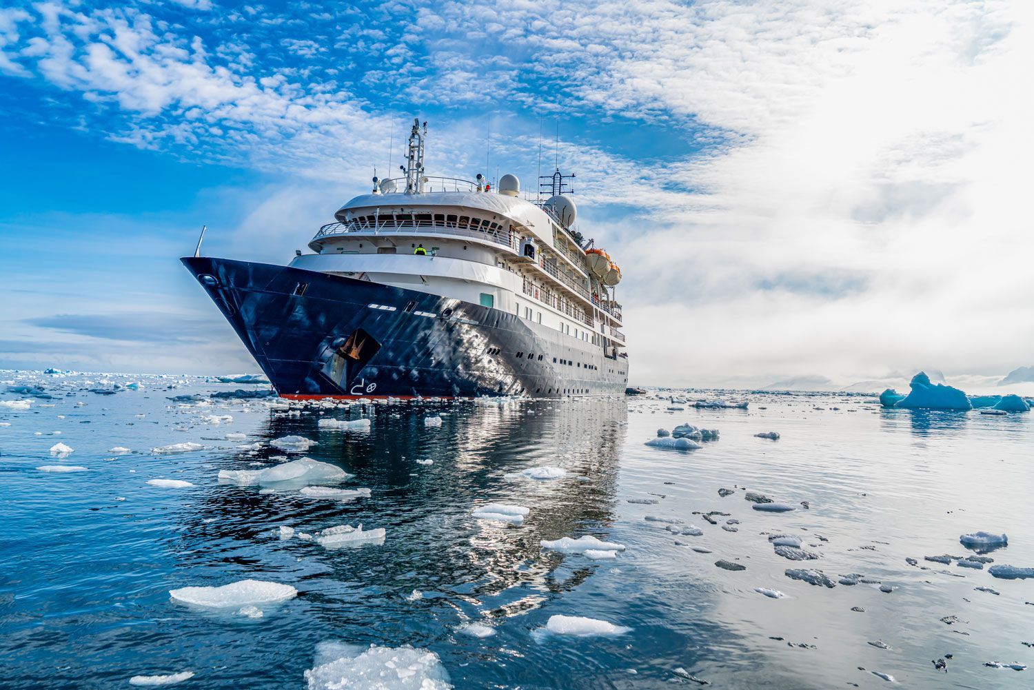 Icebreaker ship cutting through icy water under a bright blue sky, surrounded by floating icebergs.