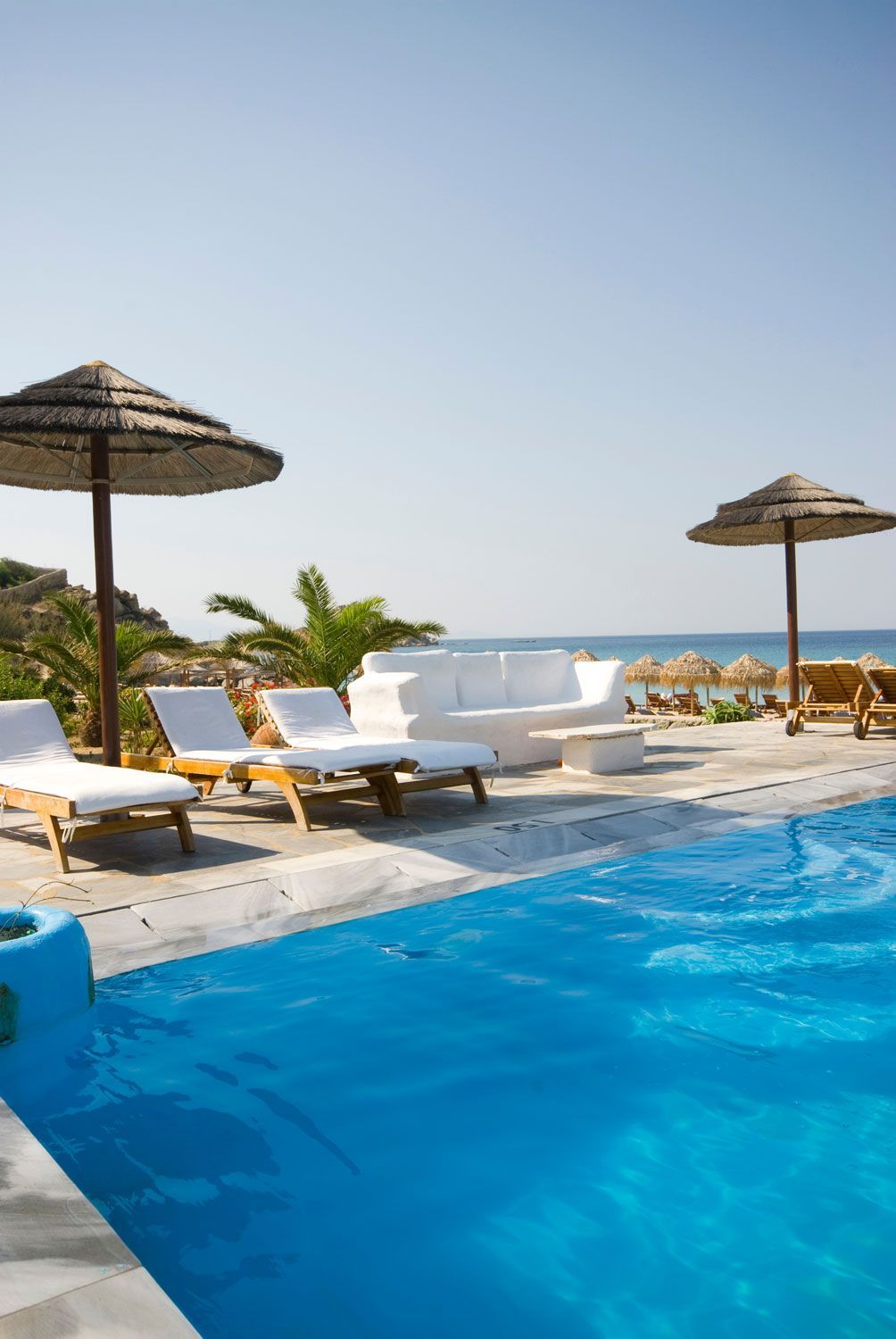 Poolside lounge chairs and umbrellas beside a bright blue resort pool with the ocean in the background
