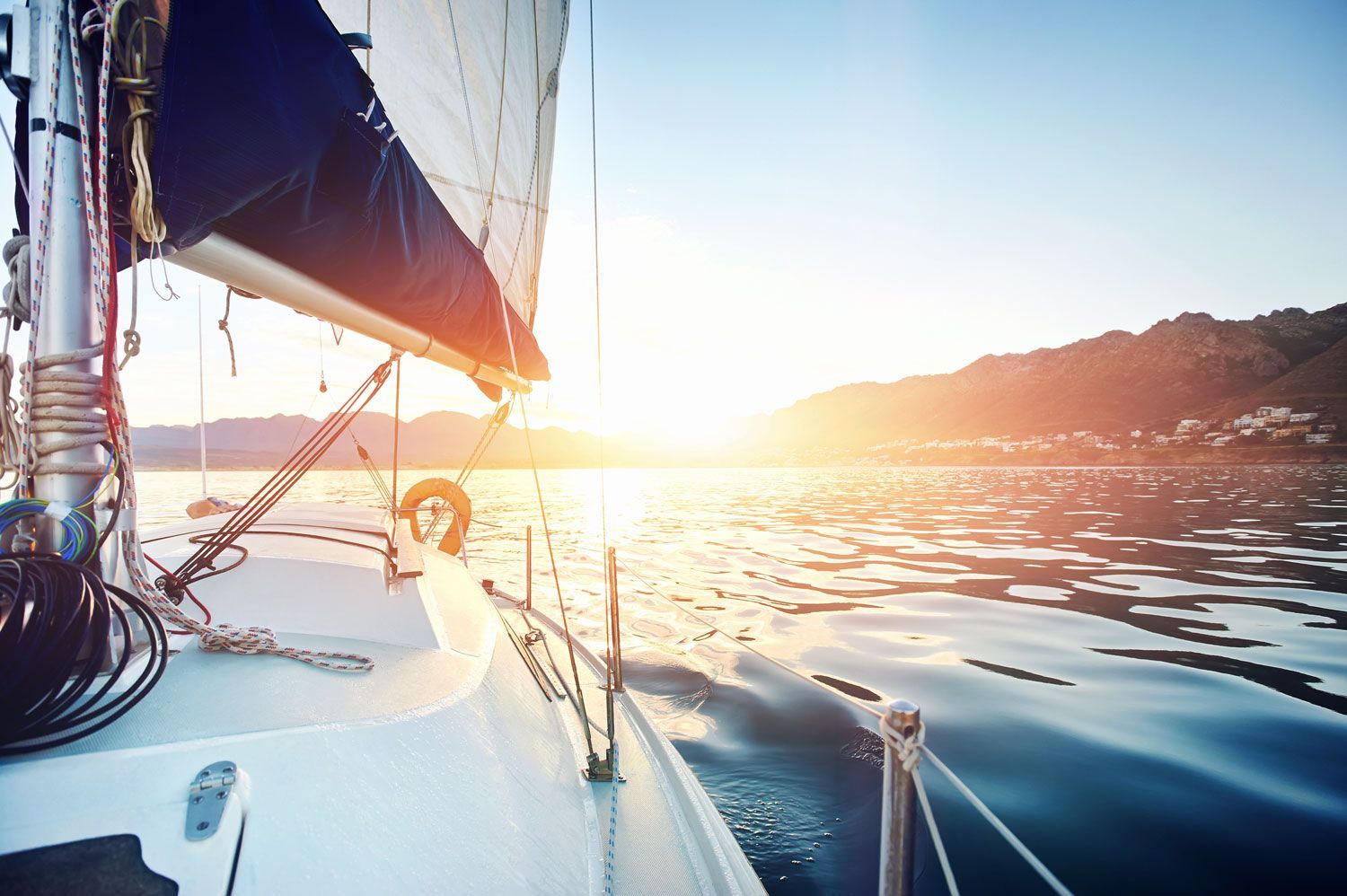 Sailboat deck at sunset on calm water, with the sail and rigging silhouetted against the glowing horizon
