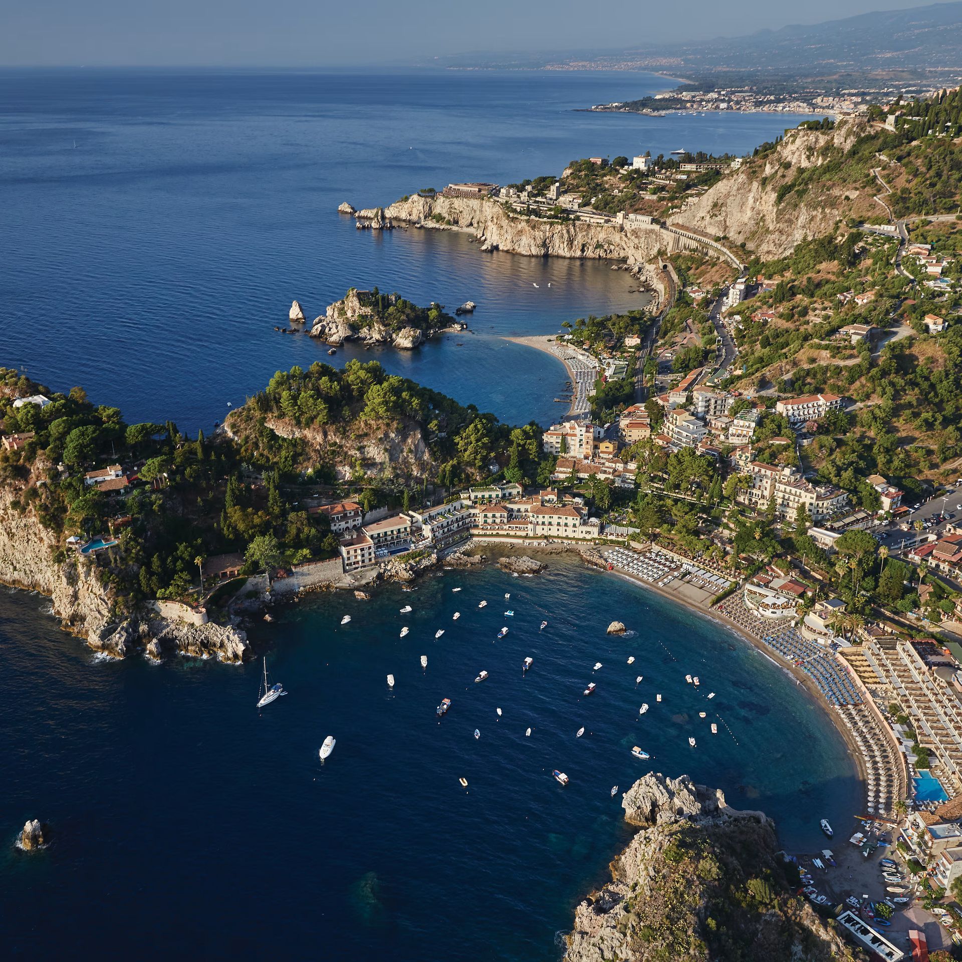Aerial view of a rocky coastal bay with blue water, boats, beaches, and hillside buildings