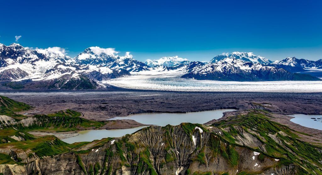 Snowy mountains rise above a wide lake and rocky green valley under a clear sky.