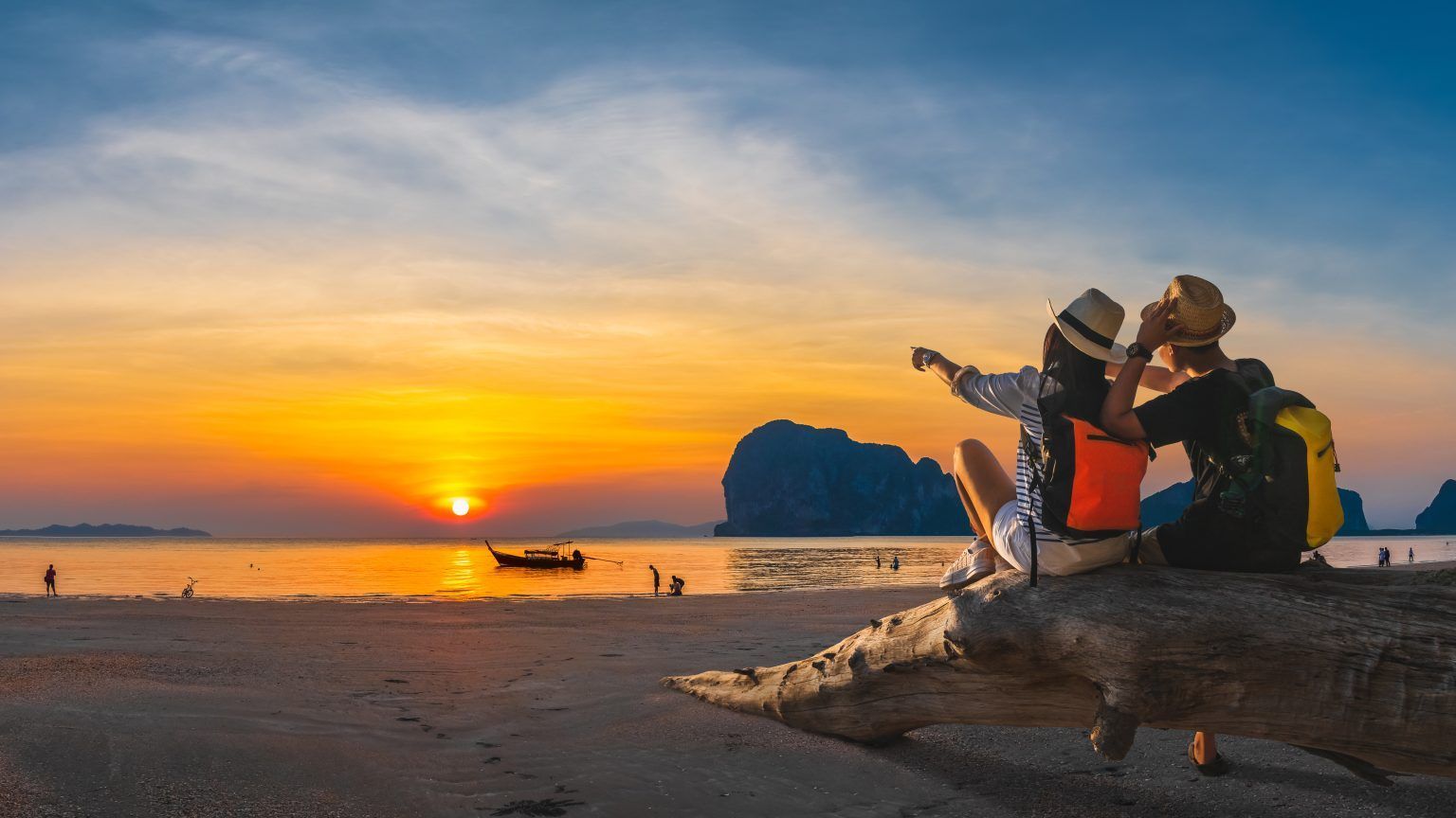 Two people sitting on a log at sunset, watching boats on a beach with a distant rock formation Two people sitting on a log at sunset, watching boats on a beach with a distant rock formation