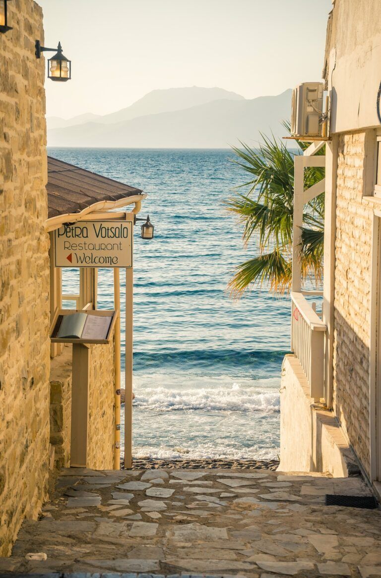 Narrow stone alley leading to a sunny beach and blue sea, with a hanging sign and palm tree at the entrance