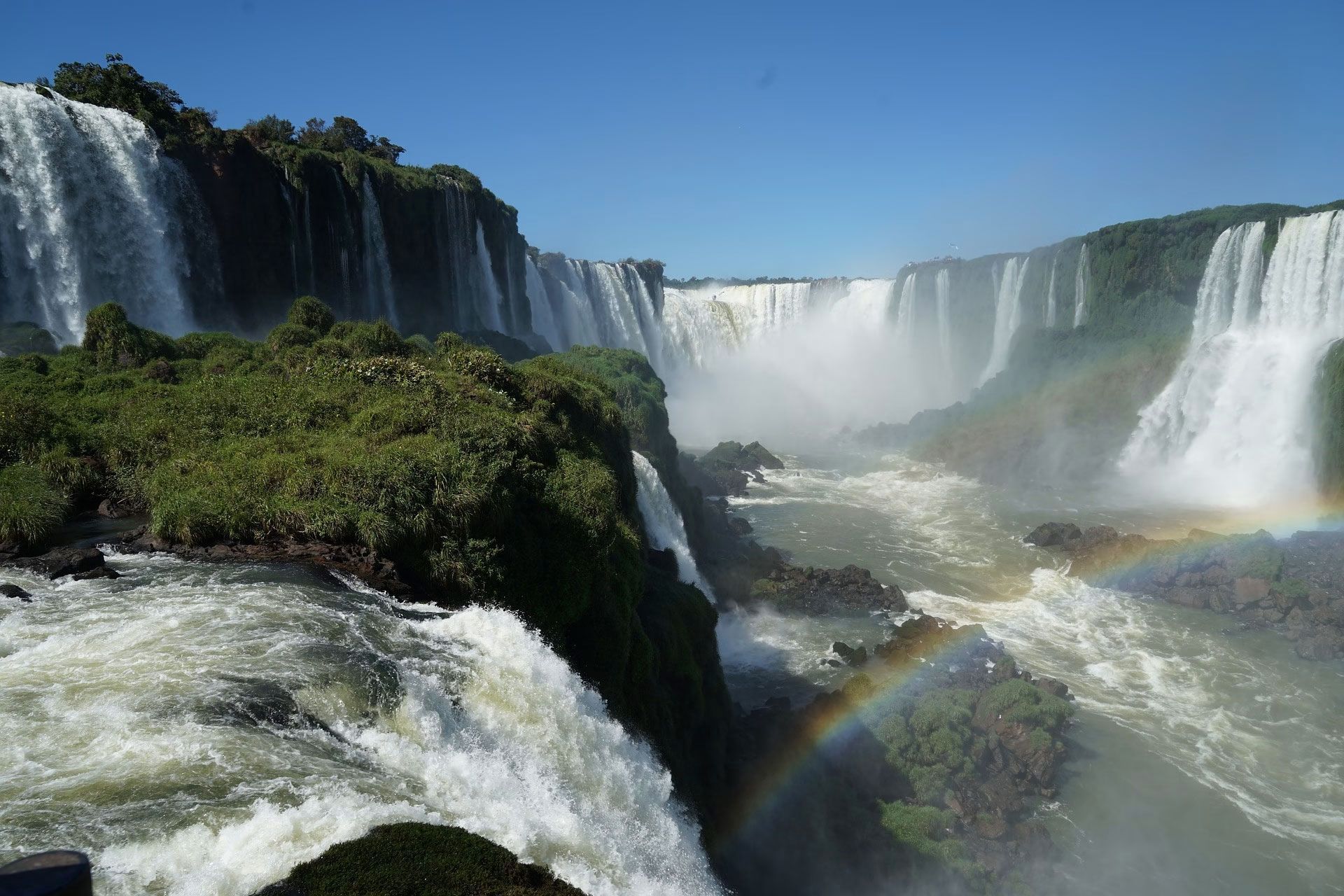Wide waterfall canyon with mist, blue sky, and a faint rainbow over rushing water