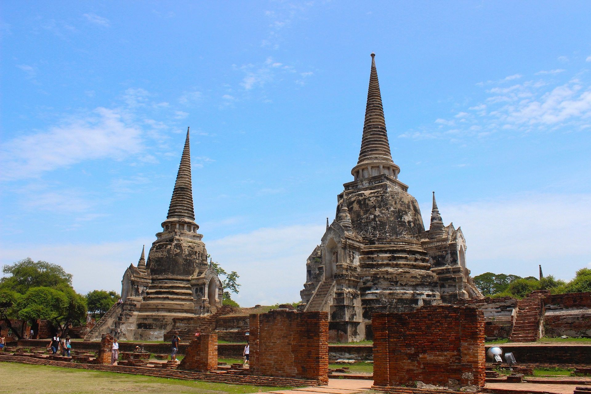 Ancient temple ruins with two stone stupas under a clear blue sky, framed by brick walls and green trees