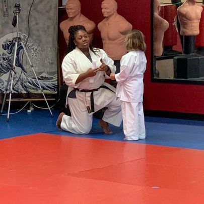 A woman in a karate uniform is kneeling down next to a little girl in a karate class.