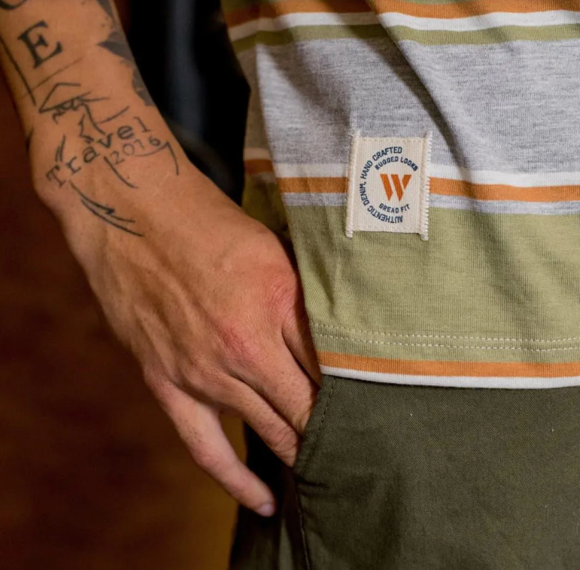 Mano sobre una camisa a rayas con una pequeña etiqueta "W" y antebrazo tatuado visible
