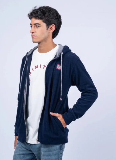 Hombre con sudadera azul marino sobre una camiseta blanca, de perfil y con las manos en los bolsillos.