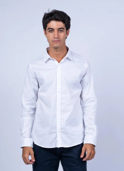 Persona de pie frente a un fondo claro, vestida con una camisa blanca y pantalones oscuros.