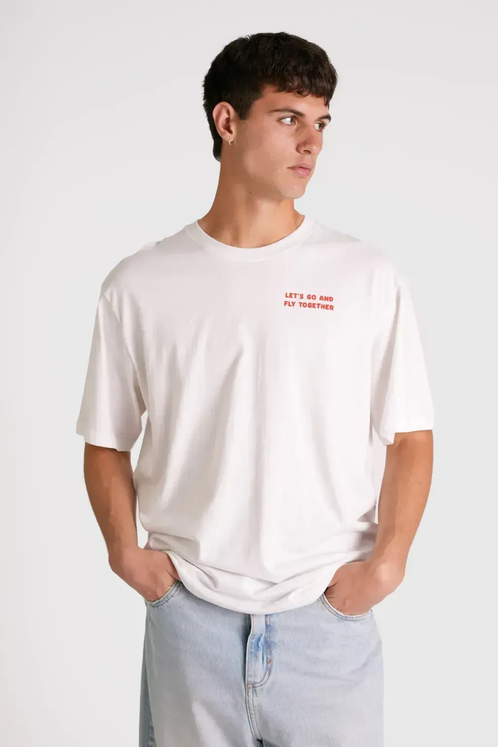 Hombre con camiseta blanca extragrande y vaqueros azul claro, de pie con las manos en los bolsillos sobre un fondo liso.