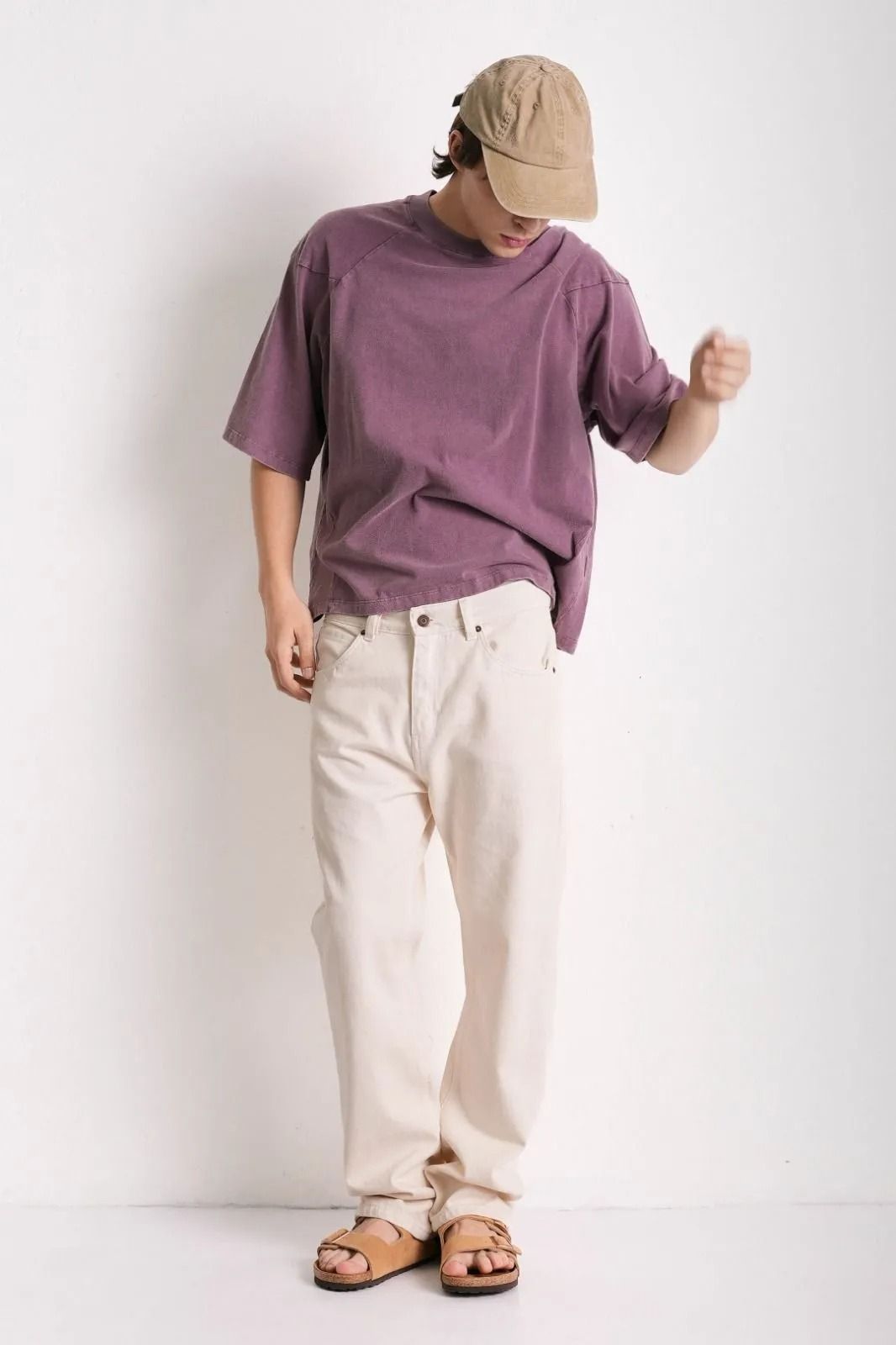 Persona con camiseta morada, pantalones color crema y sandalias, de pie sobre un fondo blanco liso.