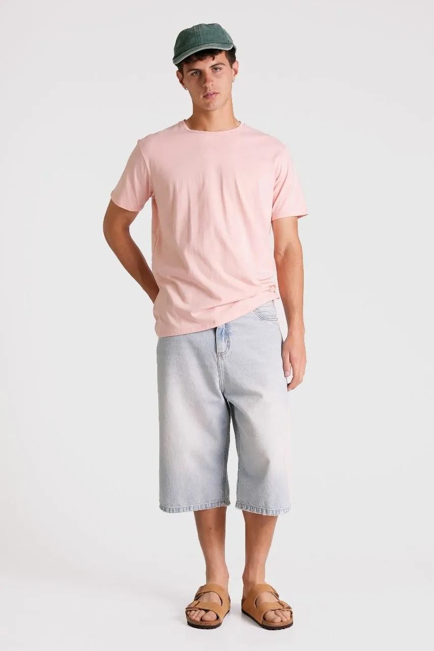 Persona con camiseta rosa, pantalones cortos gris claro, gorra verde y sandalias, de pie sobre un fondo liso.