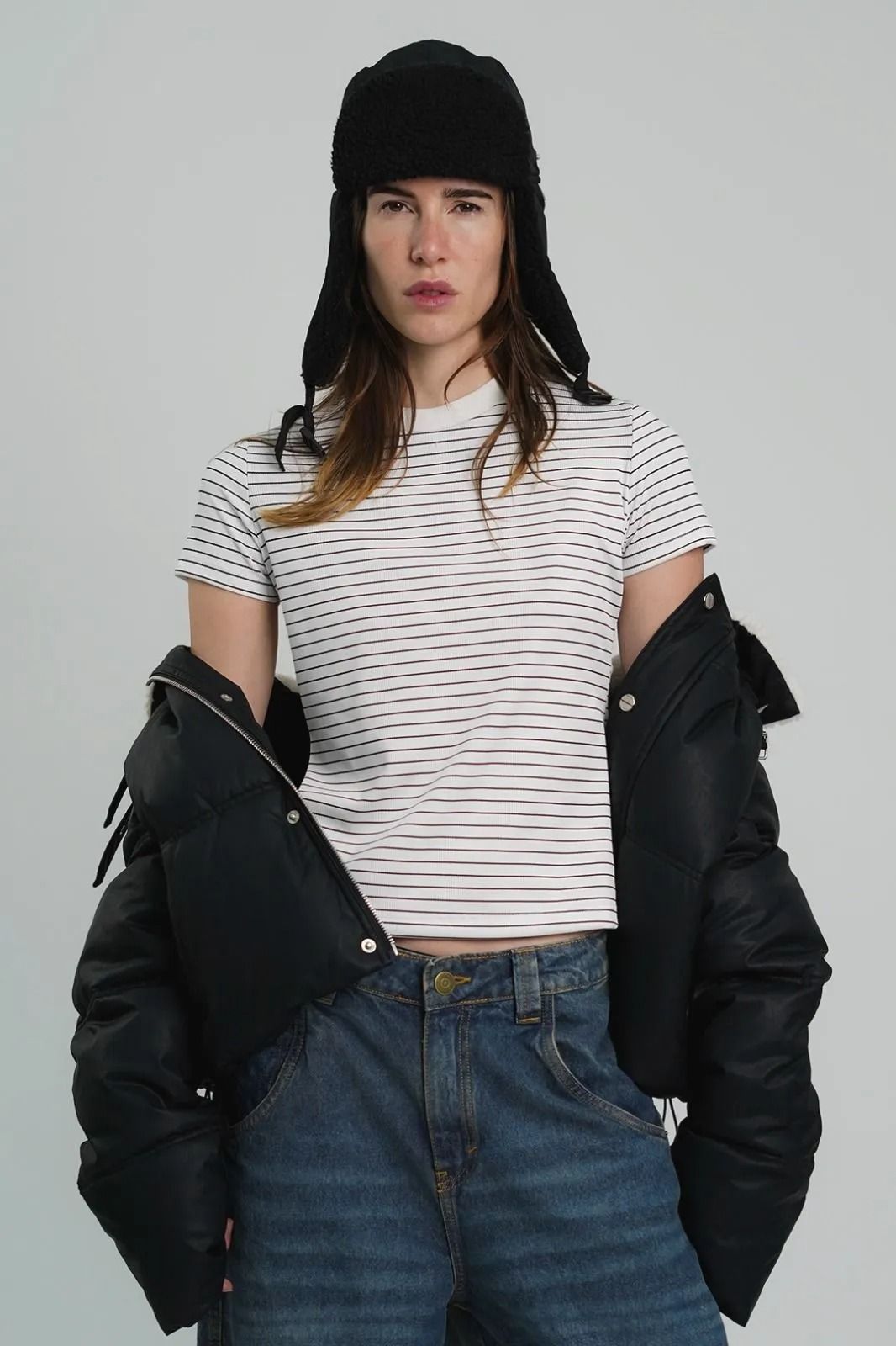 Mujer con sombrero y chaqueta negros sobre una camiseta de rayas blancas y vaqueros sobre un fondo gris.