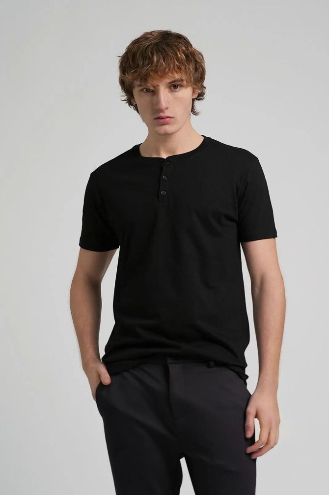 Modelo con camiseta negra y pantalones negros sobre un fondo gris claro.