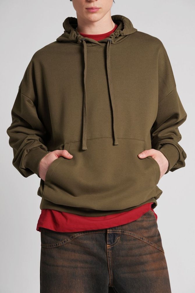 Persona con sudadera verde oliva sobre camisa roja y vaqueros, manos en los bolsillos, sobre un fondo liso.