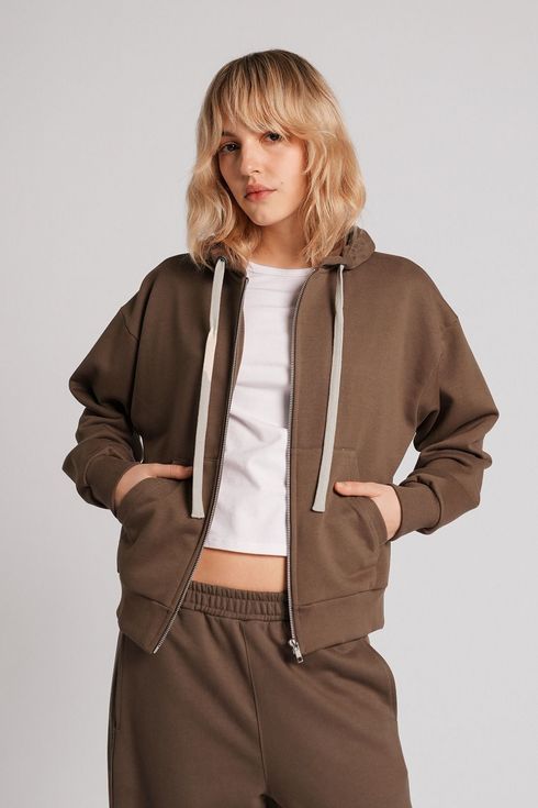 Modelo con sudadera marrón y pantalones deportivos sobre un top corto blanco, con las manos en los bolsillos, sobre un fondo gris claro.
