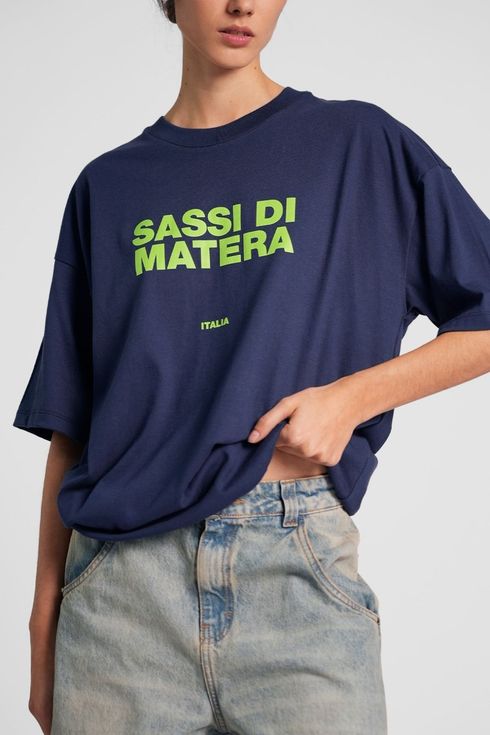 Modelo con camiseta extragrande azul marino con el texto “SASSI DI MATERA” en verde neón y vaqueros desgastados.