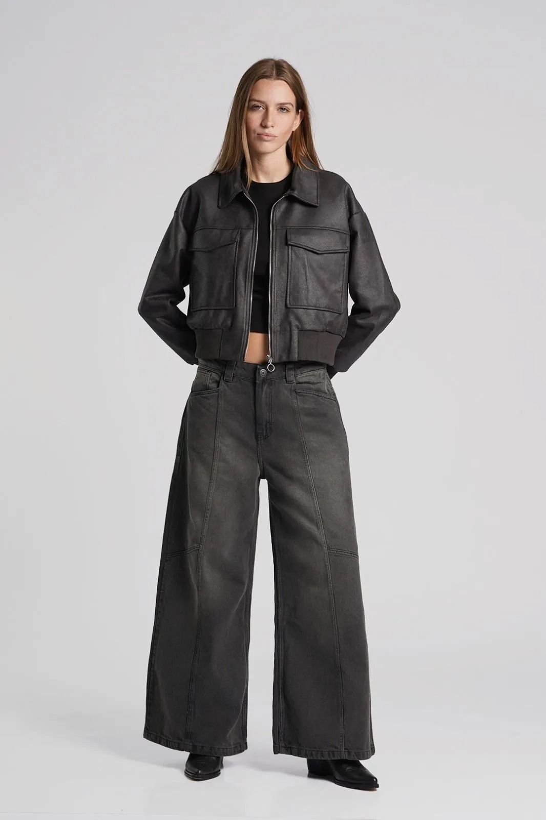 Modelo con chaqueta negra y pantalones de pierna ancha frente a un fondo de estudio gris claro.