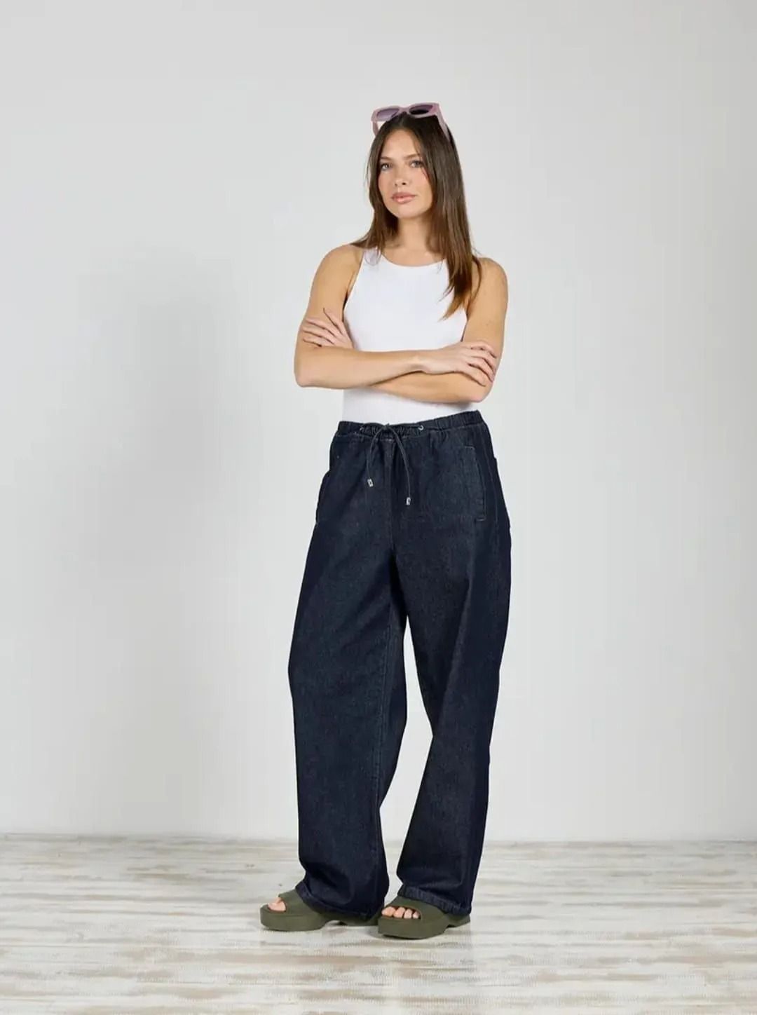 Mujer de pie con los brazos cruzados, vestida con una camiseta blanca de tirantes y vaqueros oscuros holgados, apoyada contra una pared gris lisa.