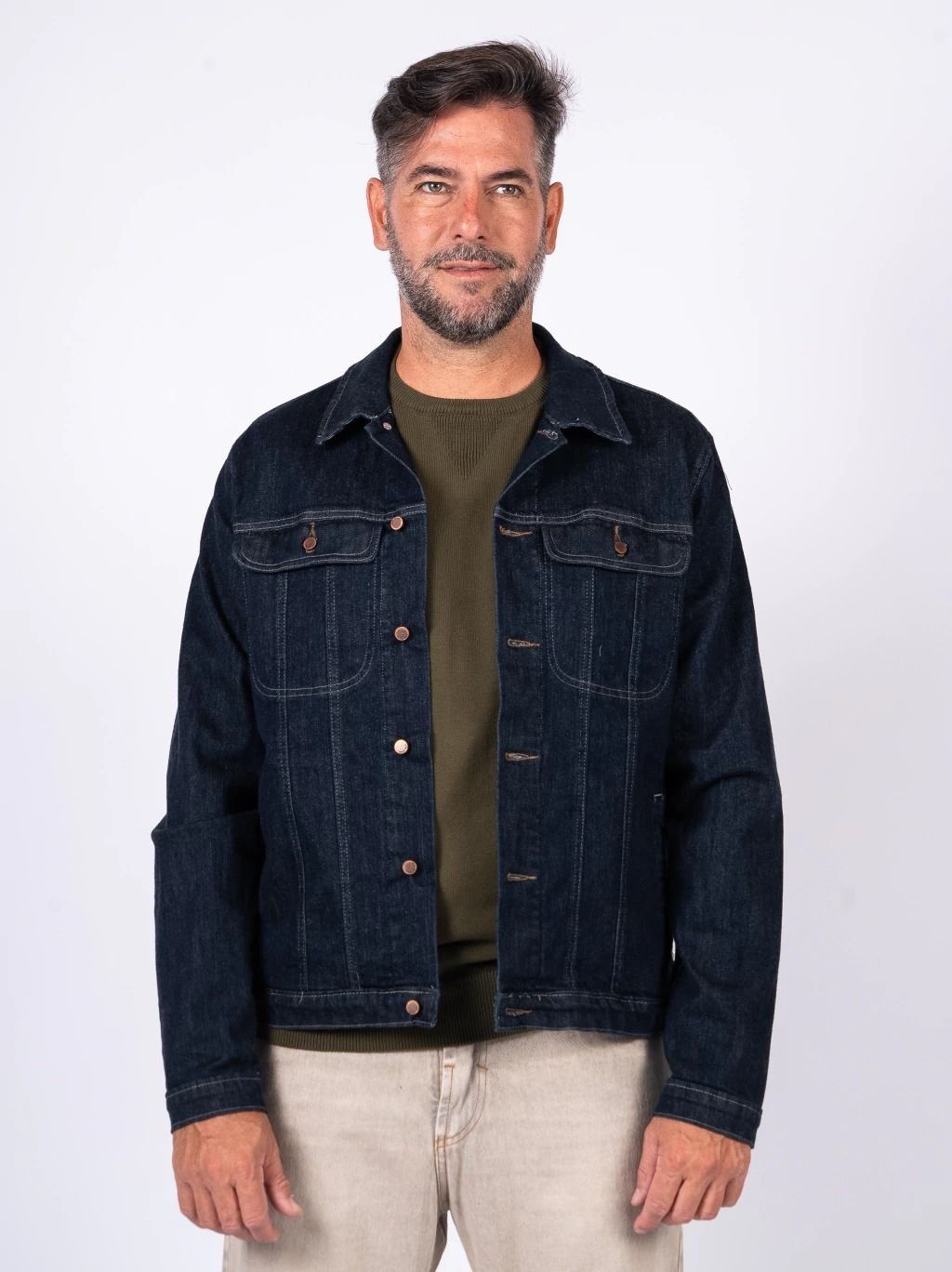 Hombre de pie sobre un fondo liso, vistiendo una chaqueta vaquera oscura, camisa verde oliva y pantalones claros.