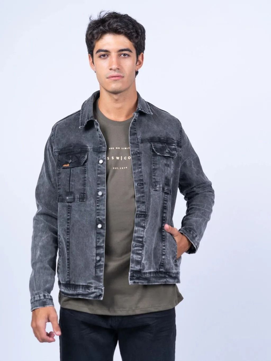 Hombre de pie sobre un fondo claro, vistiendo una chaqueta vaquera gris sobre una camiseta verde oliva y pantalones vaqueros oscuros.