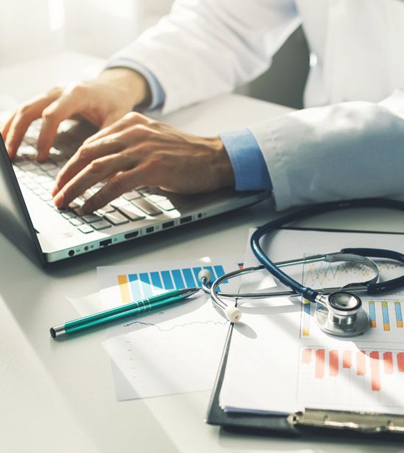 Doctor typing on laptop beside stethoscope, clipboard, and medical charts on a desk.