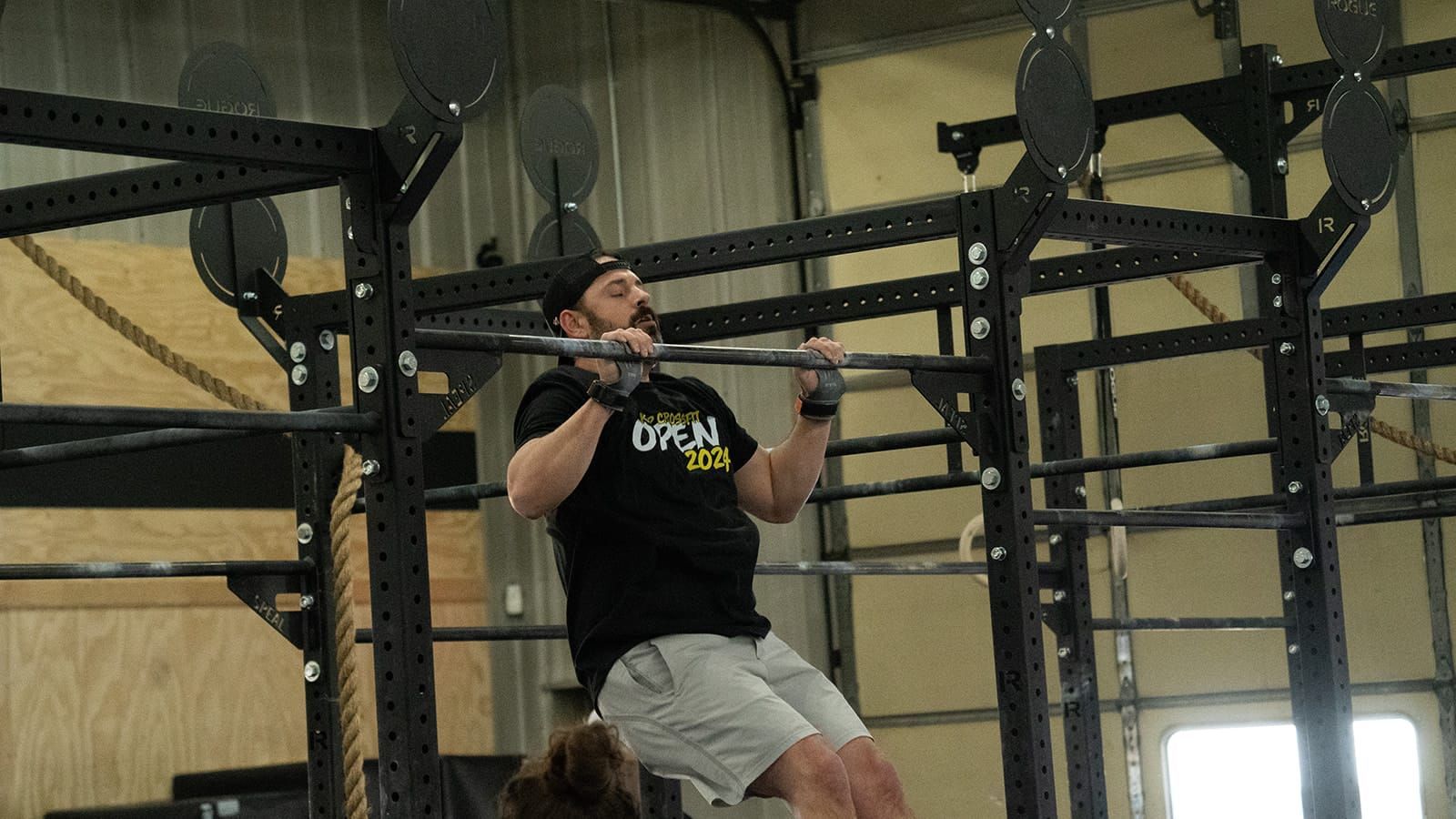 Man doing a pull-up on a black rig at a gym. He wears a black shirt, grey shorts.