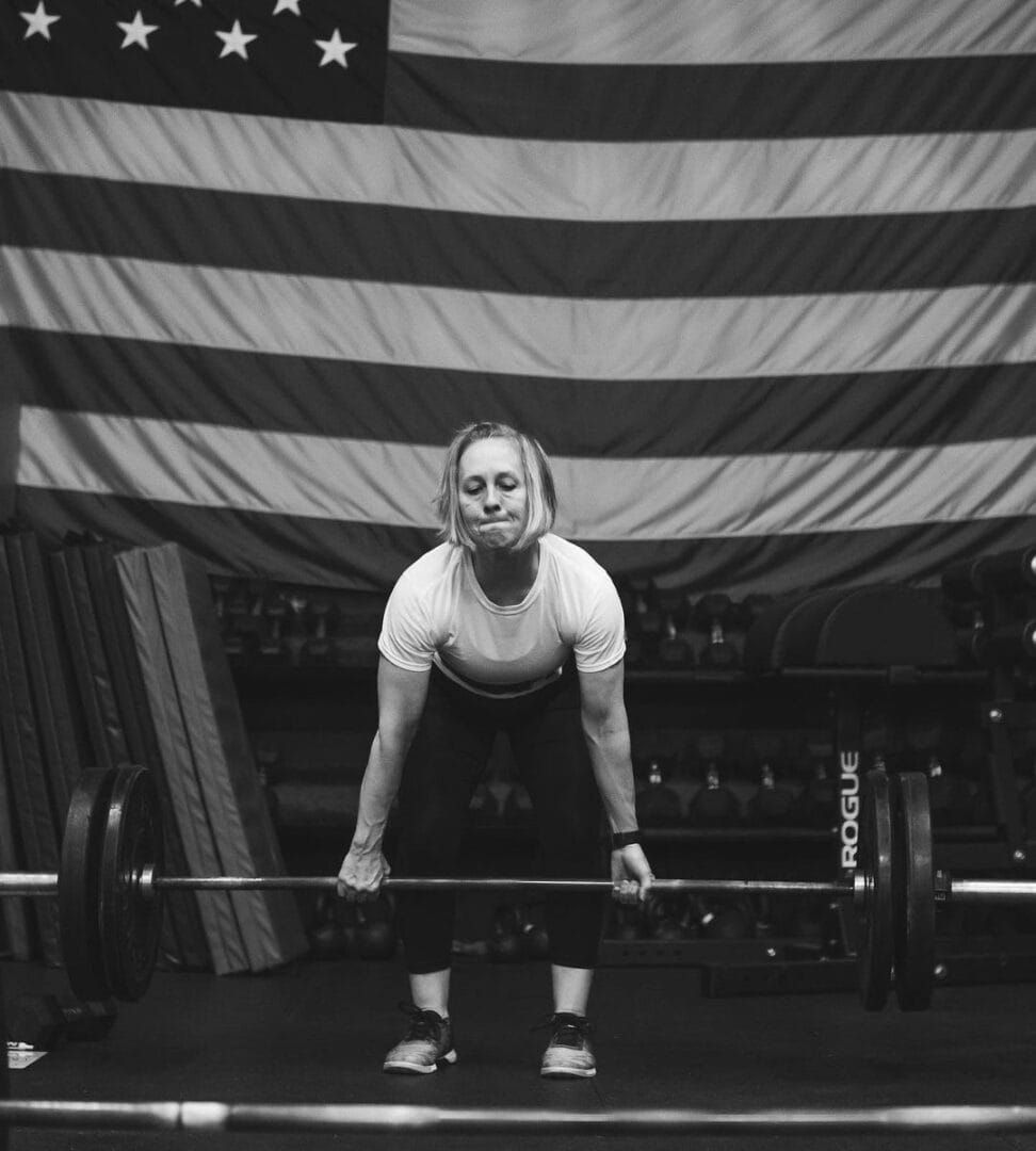 Woman in a gym lifting a barbell, American flag background.
