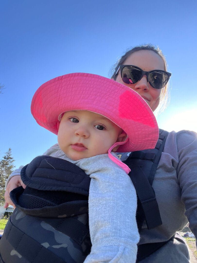 Woman in sunglasses carrying baby in a carrier, both outside on a sunny day. Baby wears a pink sun hat.