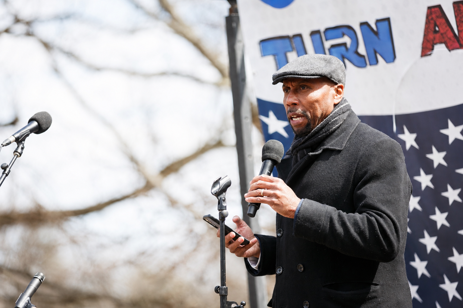 Man speaking into a microphone at an outdoor rally beside a banner with stars and partial “TURN” text
