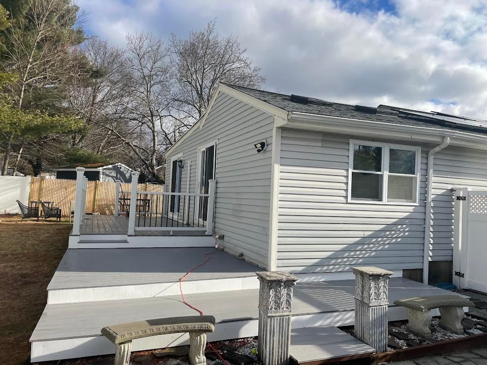 A light gray house with a small raised wooden deck, white railing, and decorative stone pillars with benches in the yard.