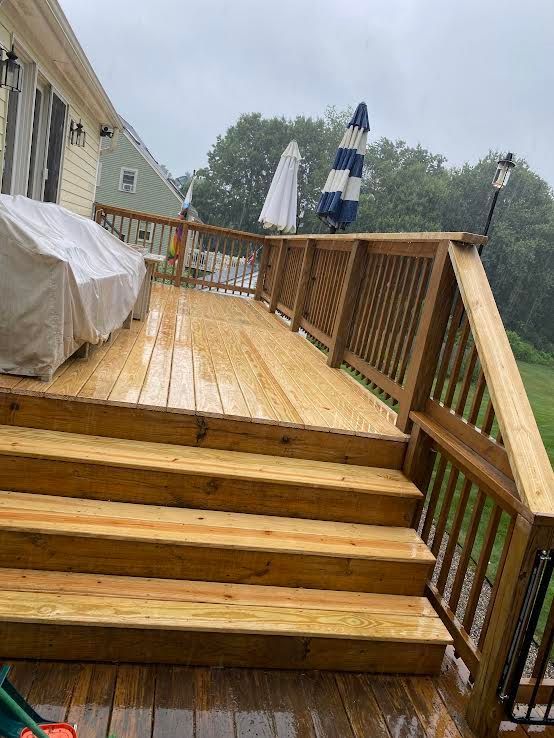 A wooden deck with stairs in the rain, featuring a covered grill and two patio umbrellas against a house exterior.