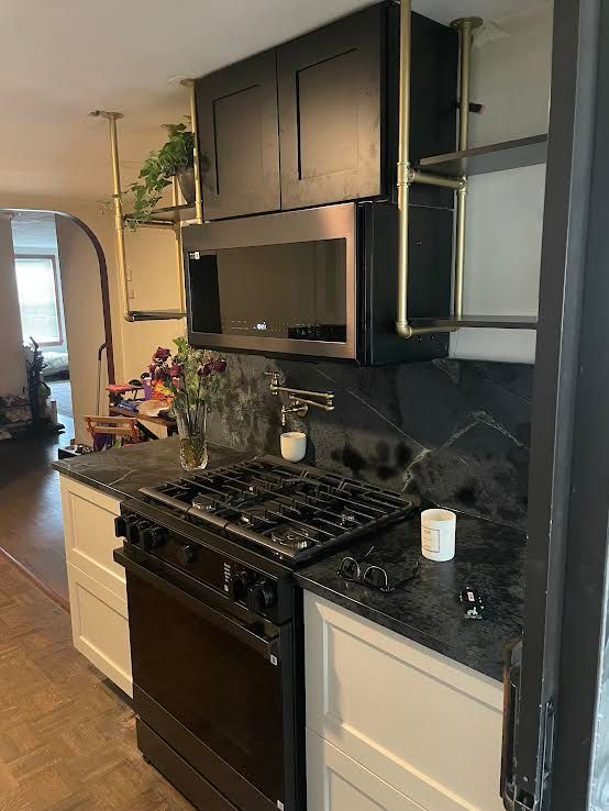 A kitchen featuring a black stove, microwave, and dark countertops, contrasted by white cabinets and gold pipe shelving.