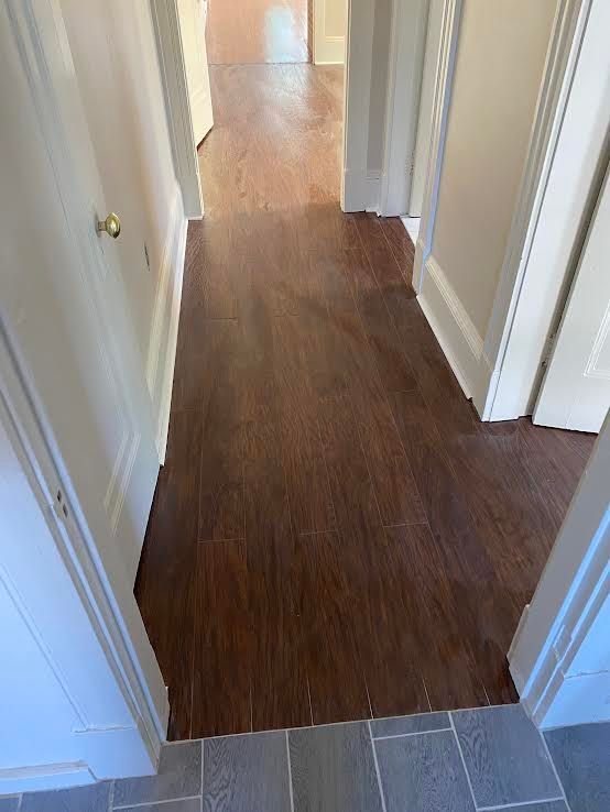 A dark wood-look hallway floor transitions to gray tile in the foreground, with white walls and door frames on both sides.