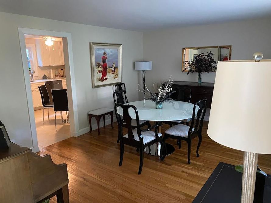 A dining room with a glass-topped table and four black chairs, hardwood floors, framed art, and a glimpse of a kitchen.