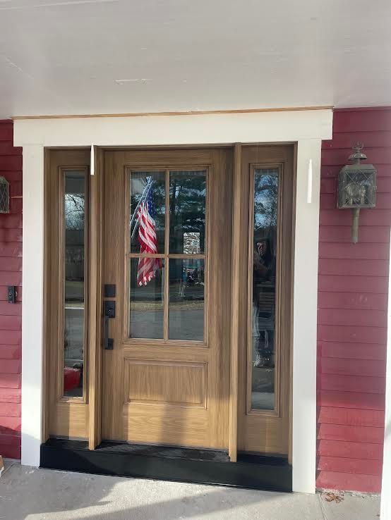 A wooden front door with a glass pane, side windows, and a white frame against red siding. An American flag is reflected.