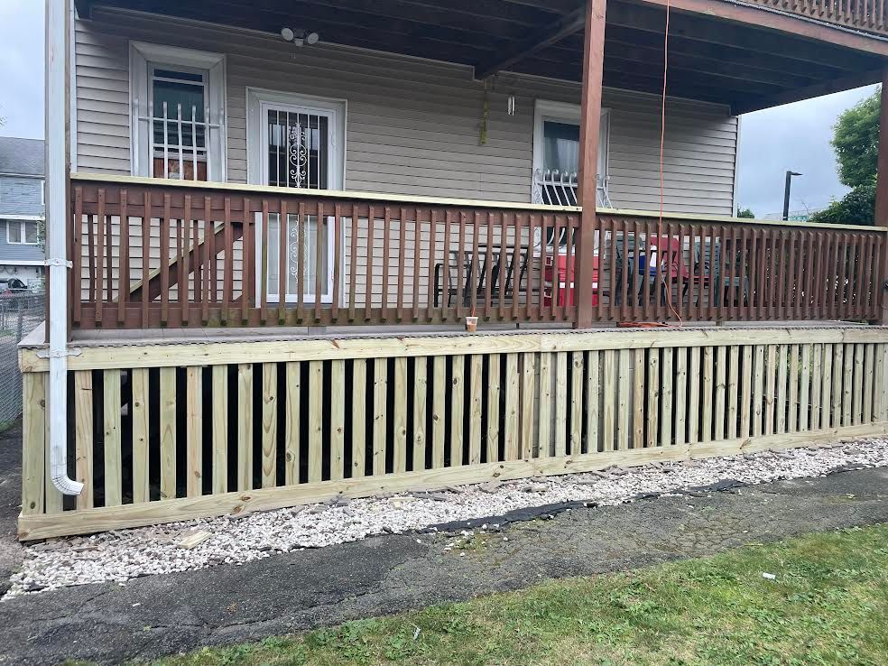 A house exterior with a brown wooden railing on a deck and new vertical pressure-treated wooden skirting along the base.