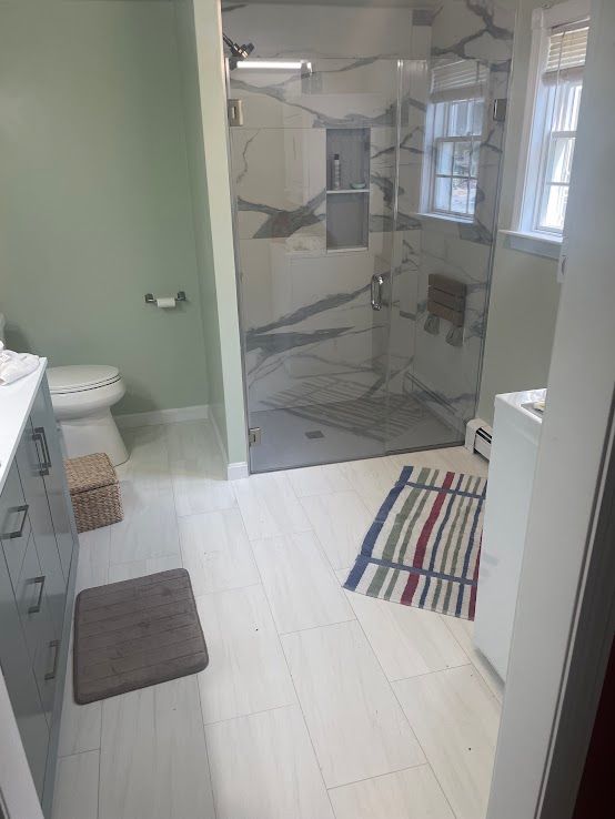 A bright bathroom with a marble-tiled walk-in shower, a grey vanity, a toilet, and striped rugs on a light-colored floor.