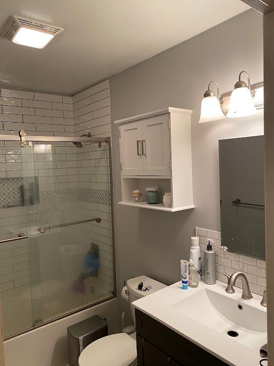 A small, bright bathroom with a white vanity, a tiled shower, a white wall-mounted cabinet, and warm vanity lighting.