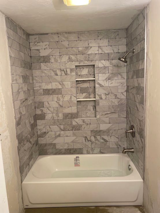 A white bathtub against a tiled wall featuring grey marble-look bricks and a recessed three-tier storage niche.