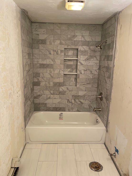 A bathroom under renovation featuring a white tub, grey marble-look tiled shower walls with a built-in niche, and floor tile.