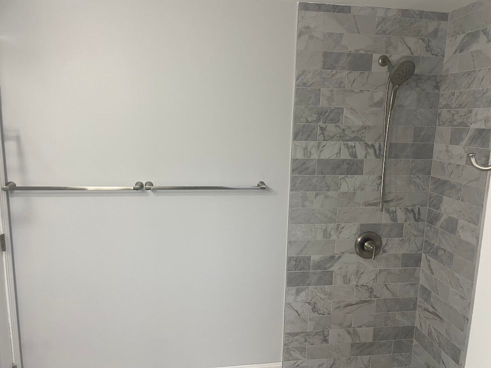A bathroom with a gray tiled shower featuring a handheld showerhead next to a light gray wall with a horizontal grab bar.