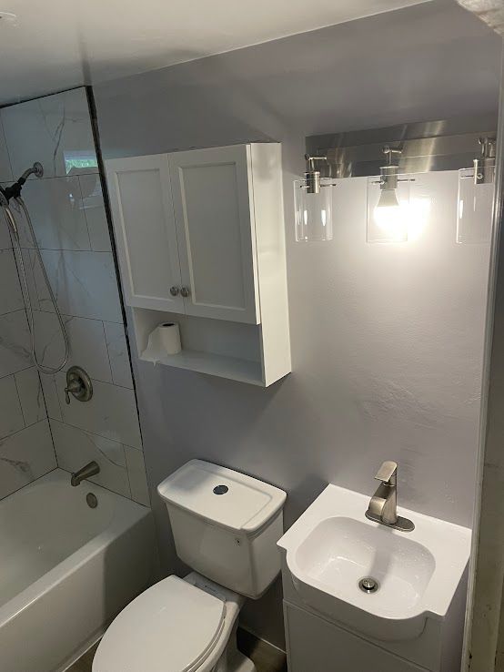 A small, modern bathroom with a white vanity sink, toilet, wall cabinet, and a bathtub with marble-tiled walls.