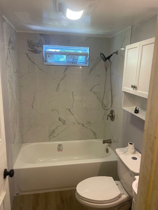 A small bathroom with a bathtub, white marble-style wall tiles, a toilet, a wooden-look floor, and a white cabinet.