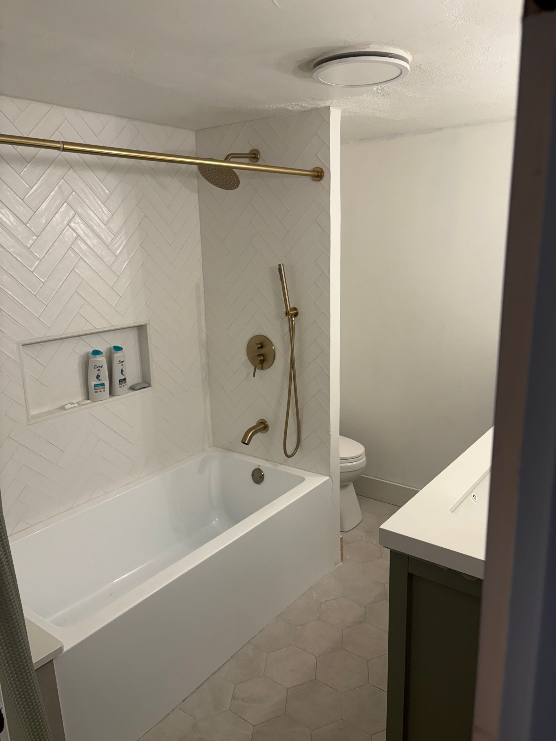 A modern bathroom featuring a white herringbone-tiled bathtub, gold fixtures, and a vanity in a small, neutral-toned space.