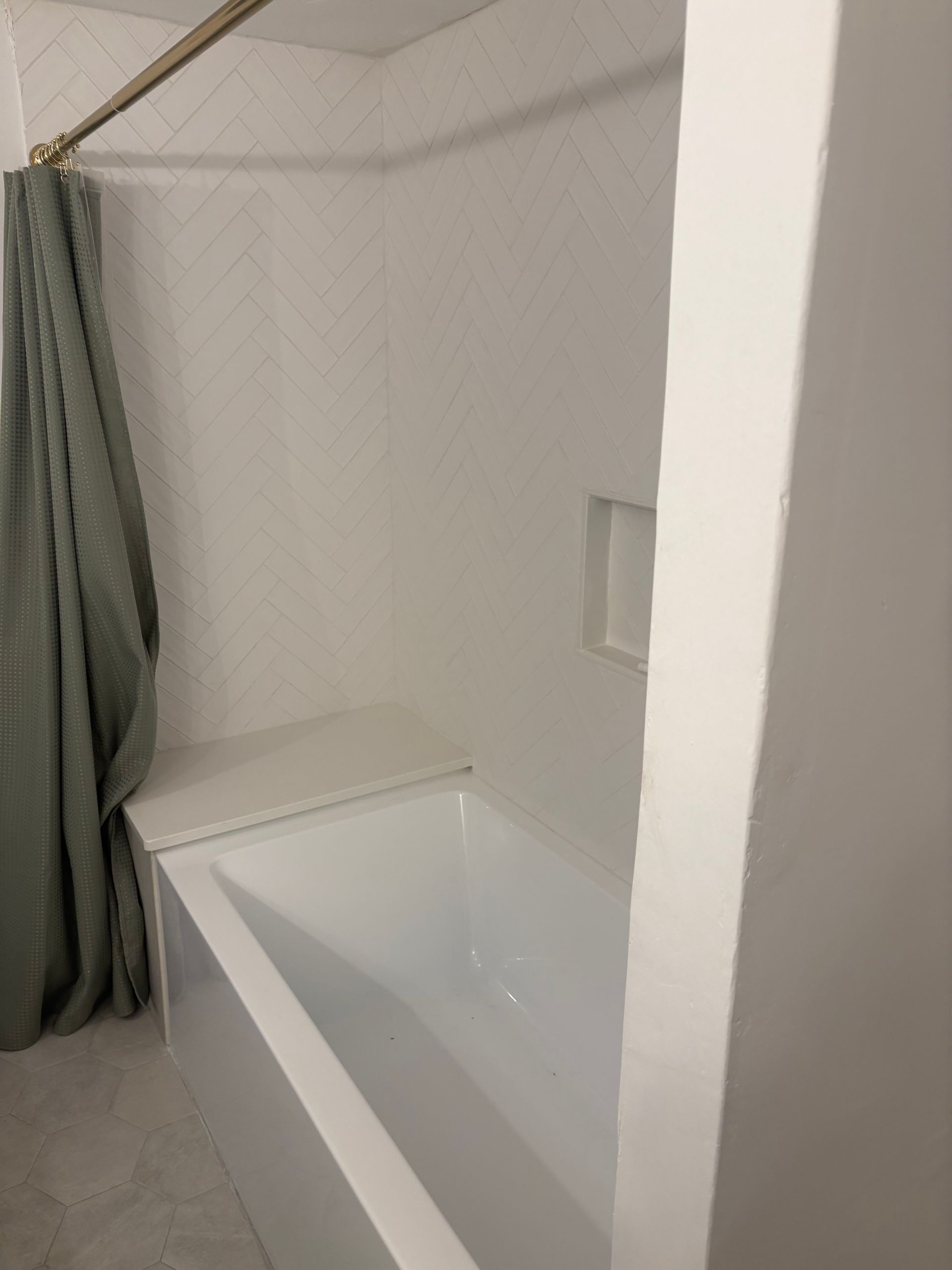 A white bathtub with a built-in seat, white herringbone tile walls, and a green shower curtain hanging on a gold rod.