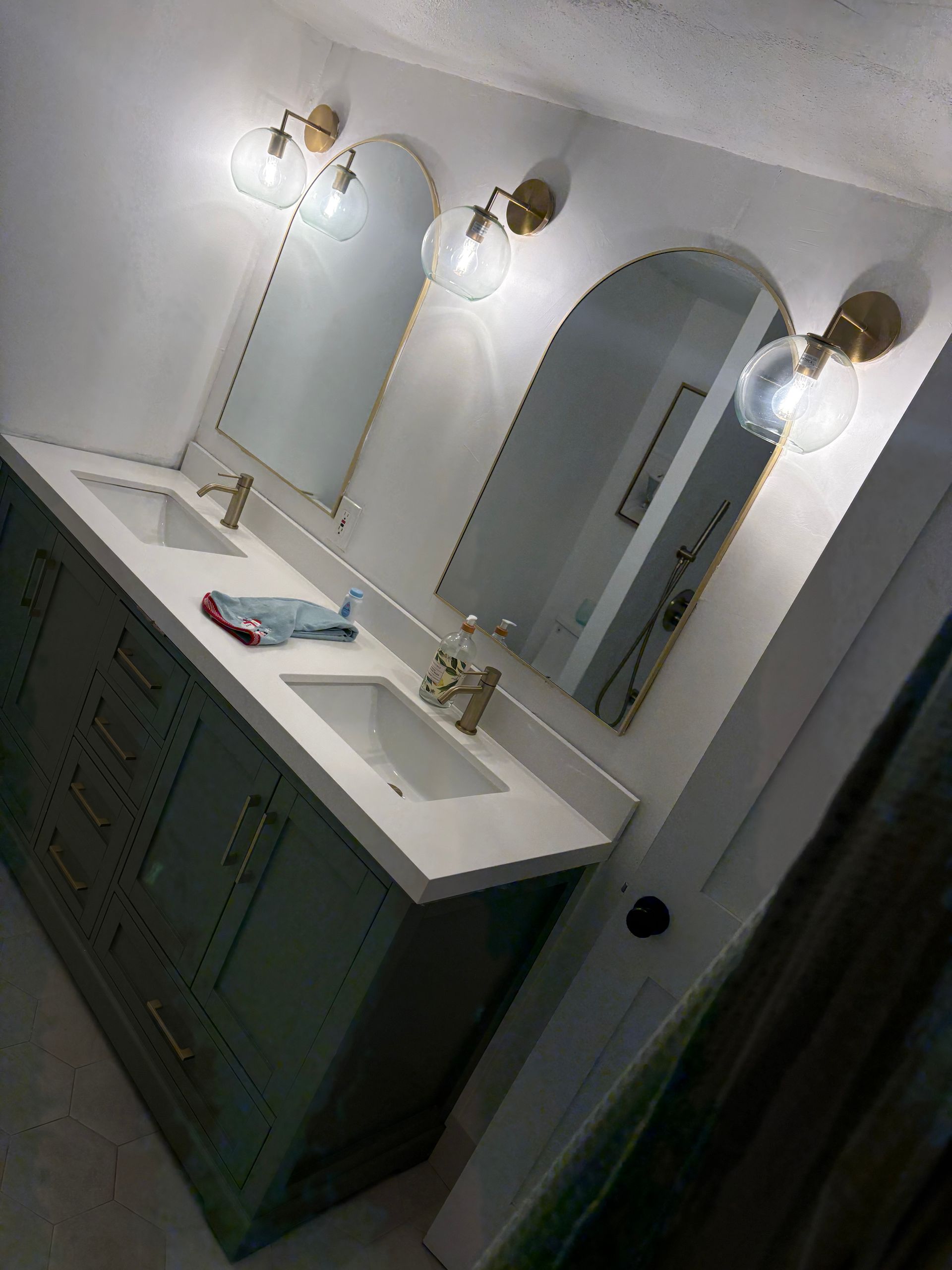 A modern bathroom vanity with a sage green cabinet, white countertop, two arched mirrors, and gold wall-mounted lights.